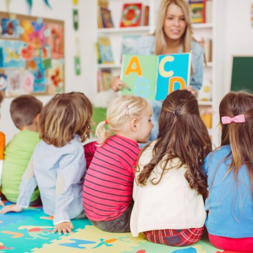 A teacher shows alphabet cards to a group of young children sitting on a colorful play mat in a classroom.